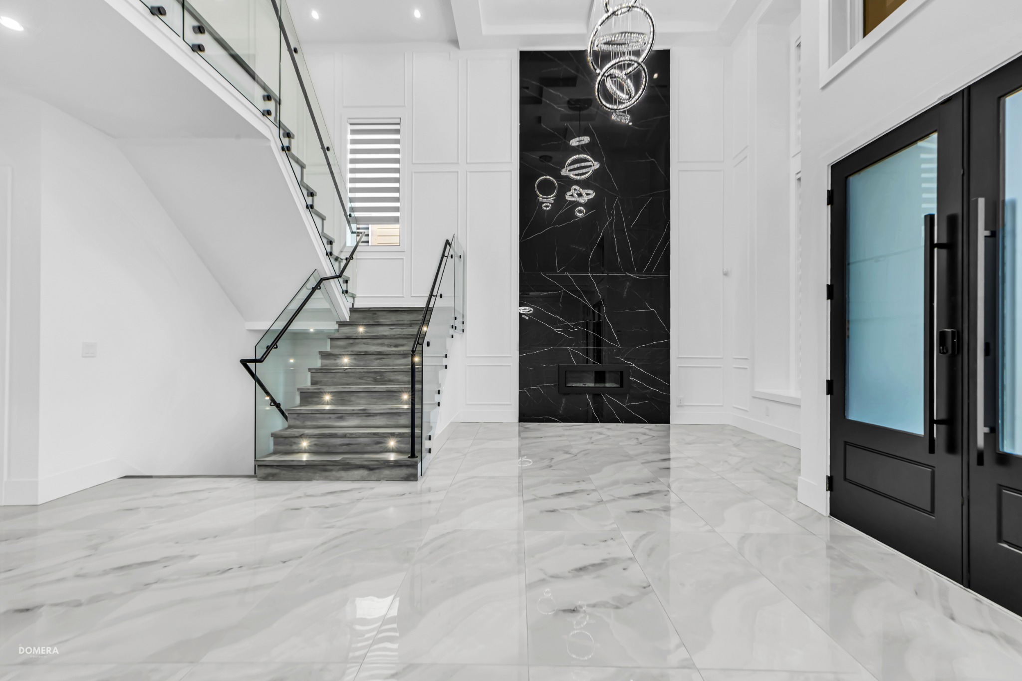 Luxury double-height entry with a glass staircase, black marble fireplace wall and sculptural ring chandelier